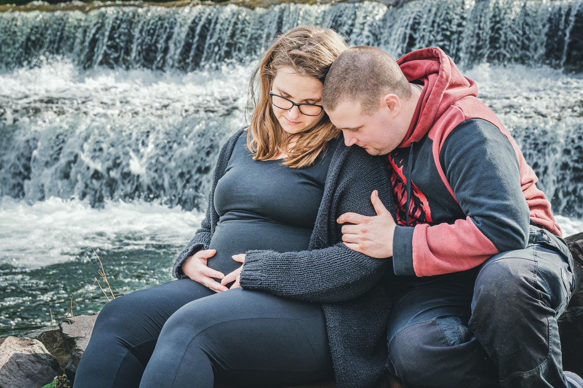 Werdende Eltern sitzten gemeinsam an einem Wasserfall und freuen sich auf ihr Baby.